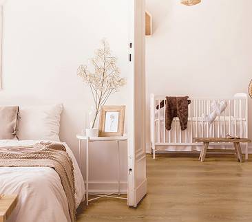 Real Photo Of White Bedroom Interior With Bedside Table With Plant, Poster And Mug And Open Door To Baby Room With Rug And White Crib